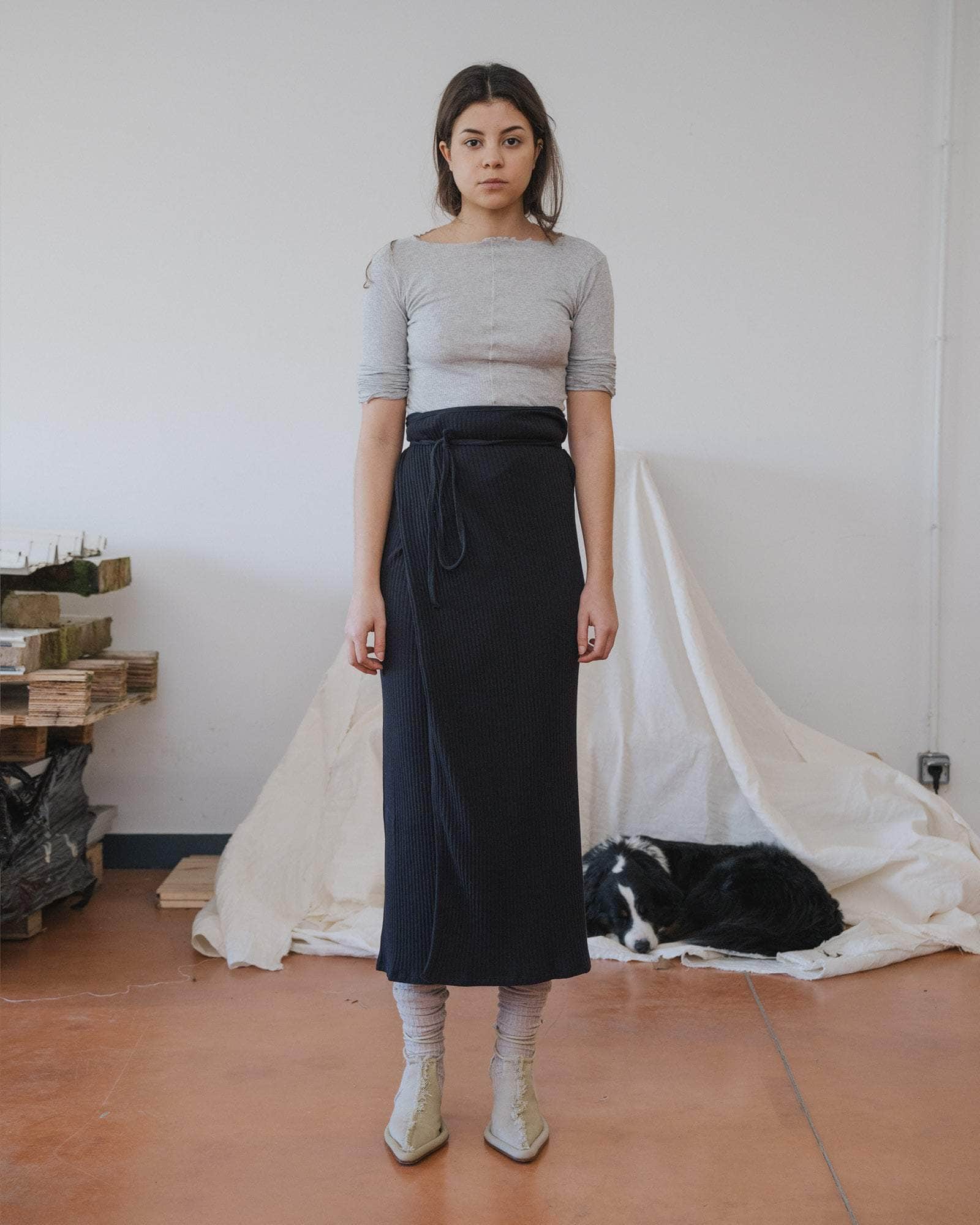 Woman wearing a gray top and black skirt standing in a room with white walls and a tiled floor.