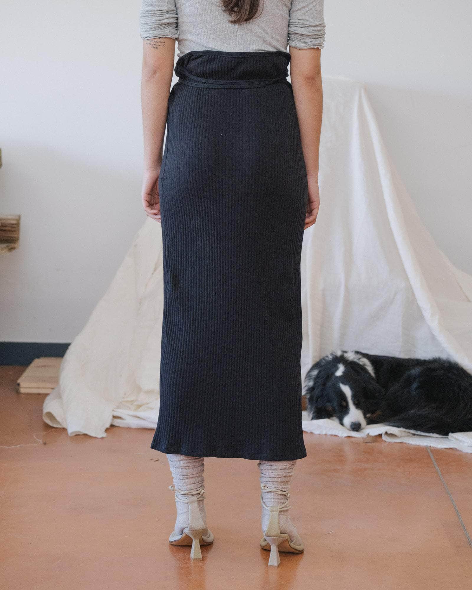 Person wearing a black ribbed skirt standing in a room with a white curtain and dog.